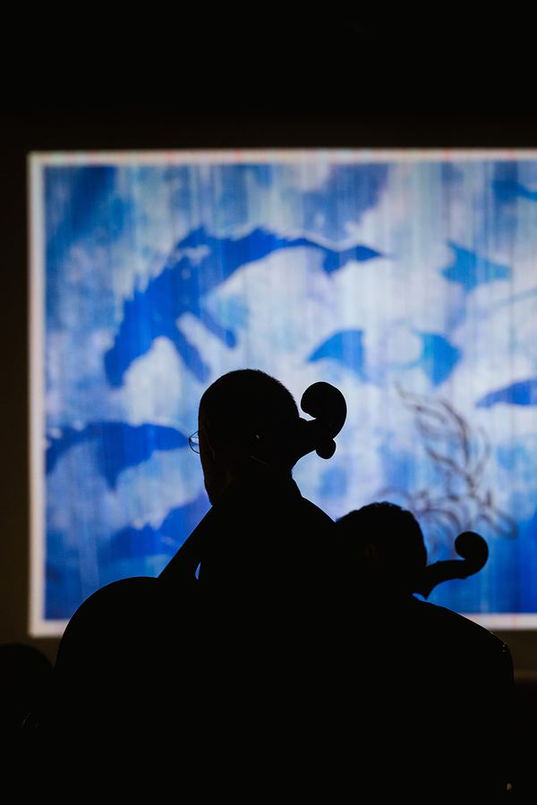Concert photo: a cellist in silhouette against a projected blue drawing
