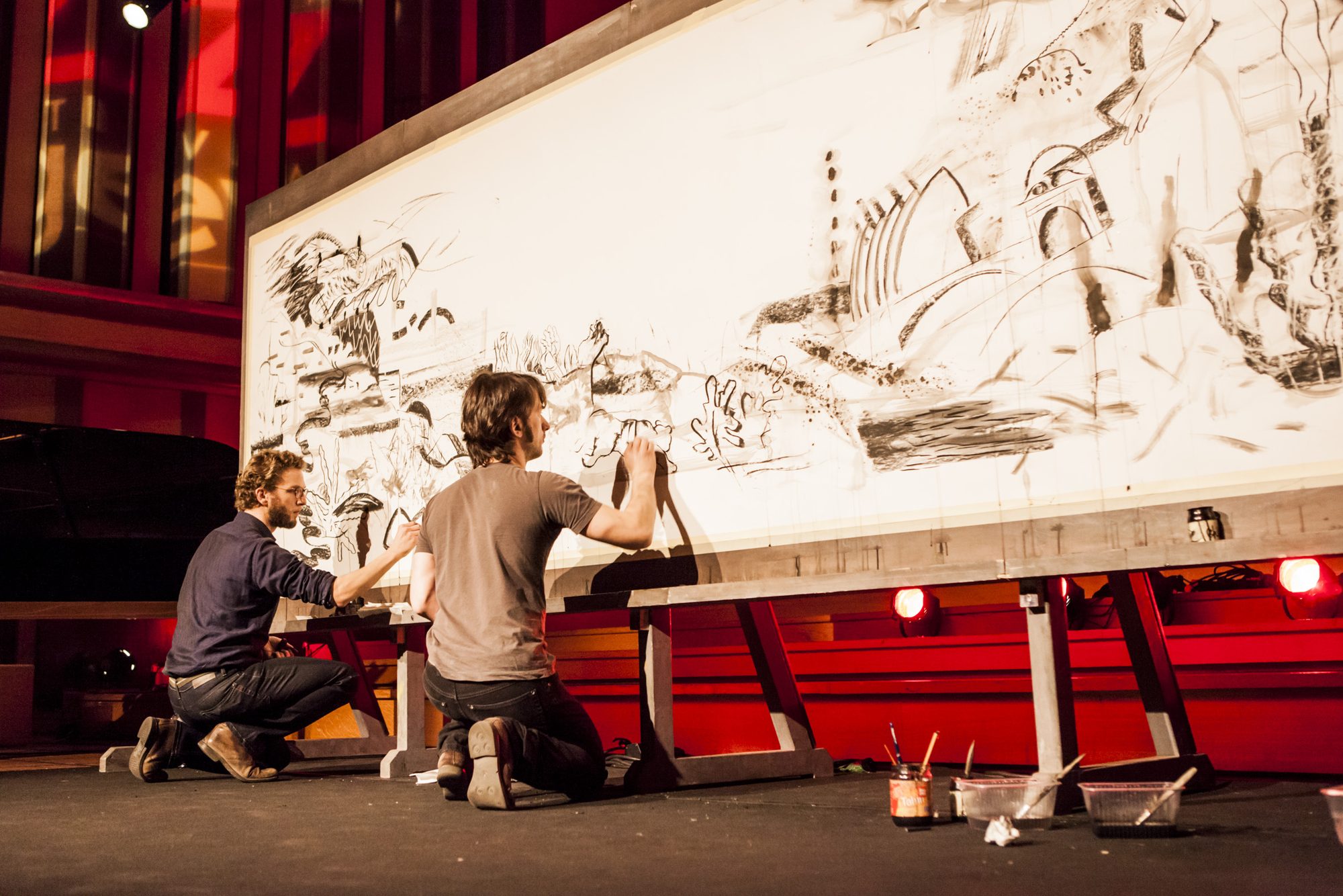 Stage photo: two artists kneeling and standing at a monumental charcoal drawing
